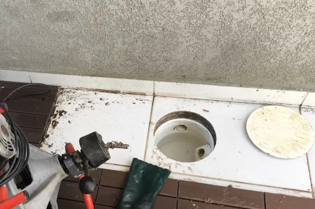 Caixa de inspeção de esgoto aberta em bancada externa, com a tampa redonda ao lado e uma máquina de desentupir