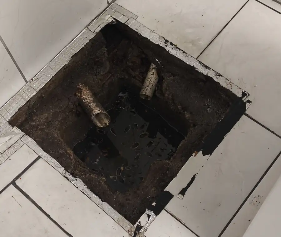 Caixa de inspeção de esgoto aberta no piso de cerâmica, com dois tubos de entrada à vista