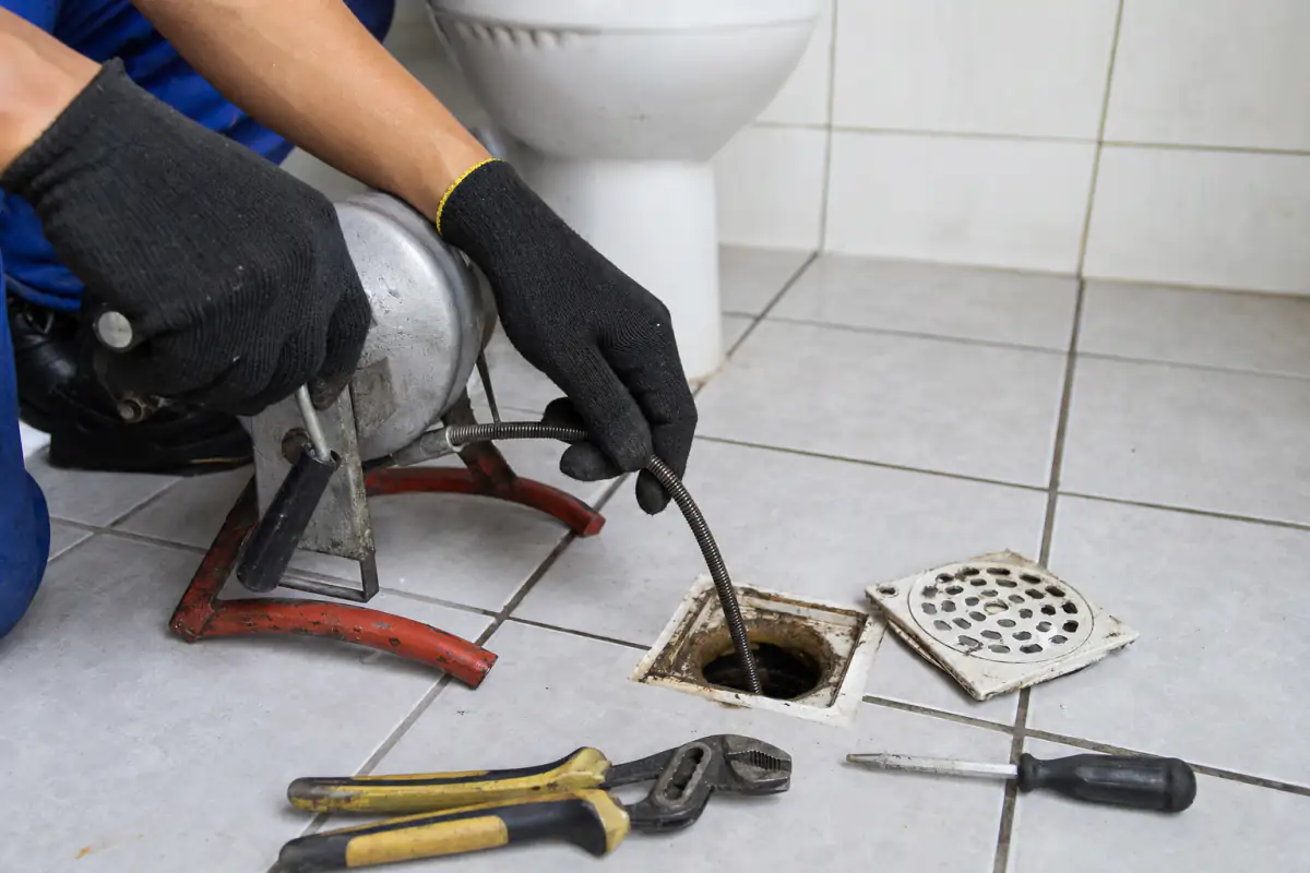 Mãos com luvas passando uma mola de desentupir com tambor e manivela por um ralo aberto de piso cerâmico
