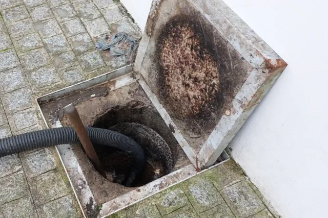 Caixa de esgoto aberta no calçamento de pedra, com mangueira preta de limpeza dentro e a tampa virada com resíduo de gordura