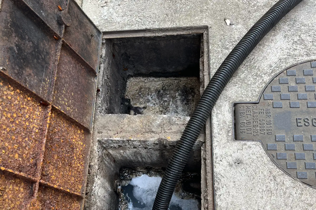 Como Desentupir Caixa de Gordura: Passo a Passo Seguro 2 Caixa de gordura de alvenaria com duas câmaras, tampa metálica enferrujada aberta à esquerda e mangueira preta entrando na câmara