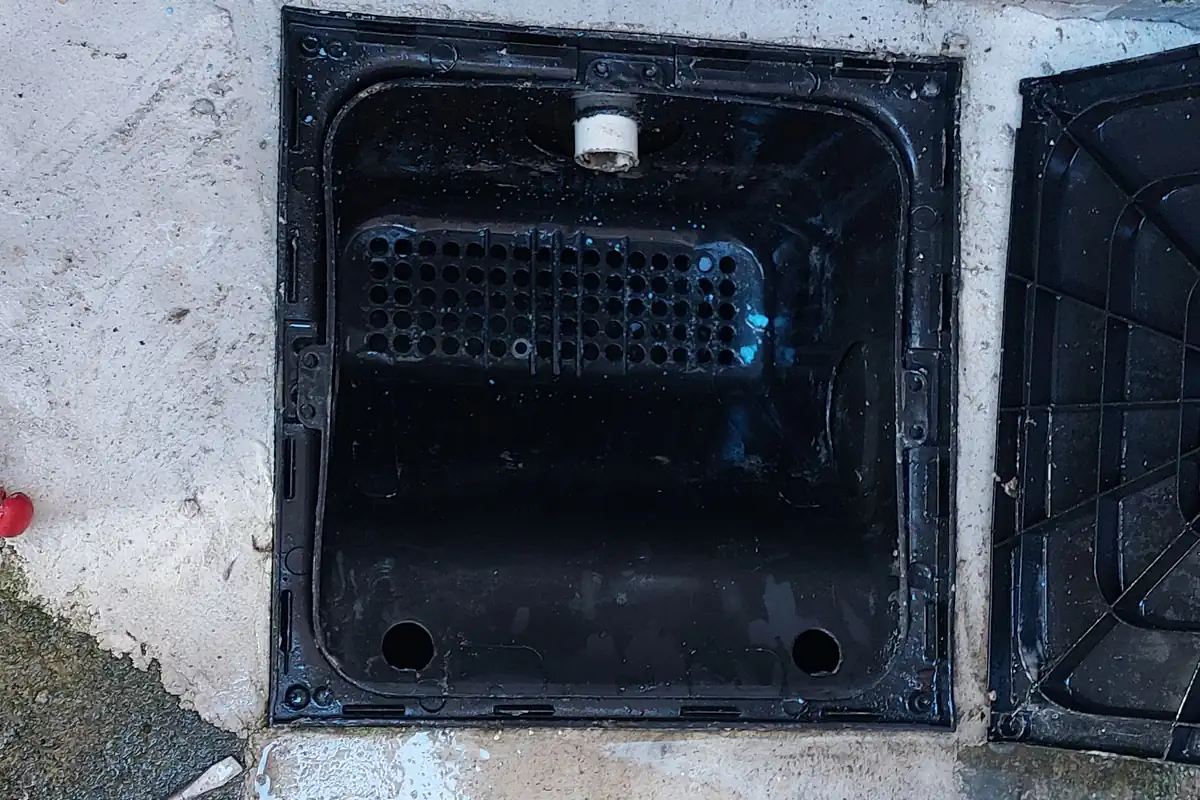 Caixa de Gordura Entupida: O Que Fazer e Quando Chamar 3 Caixa de gordura de PVC preta, limpa, aberta no piso, com o cesto de limpeza visível e a tampa ao lado