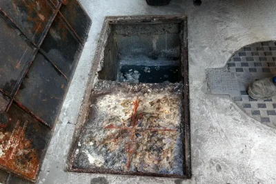 Caixa de gordura retangular aberta no piso de concreto, com a metade da frente coberta por gordura e resíduos brancos e alaranjados