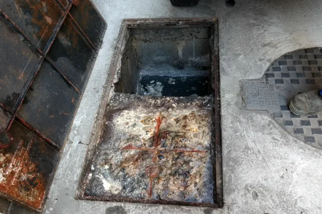 Caixa de gordura retangular aberta no piso de concreto, com a metade da frente coberta por gordura e resíduos brancos e alaranjados