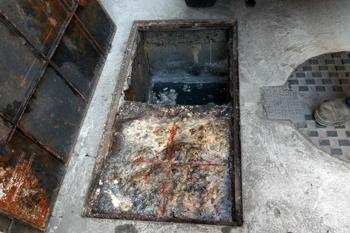 Caixa de gordura retangular aberta no piso de concreto, com a metade da frente coberta por gordura e resíduos brancos e alaranjados