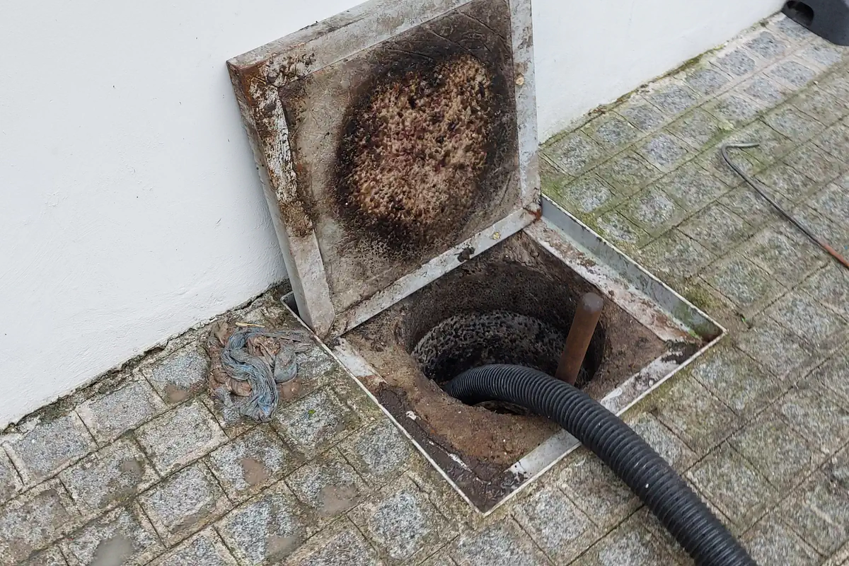 Como Desentupir Caixa de Gordura: Passo a Passo Seguro 1 Caixa de gordura quadrada no piso de pedras de uma calçada, com a tampa aberta coberta de gordura escura e uma mangueira preta dentro