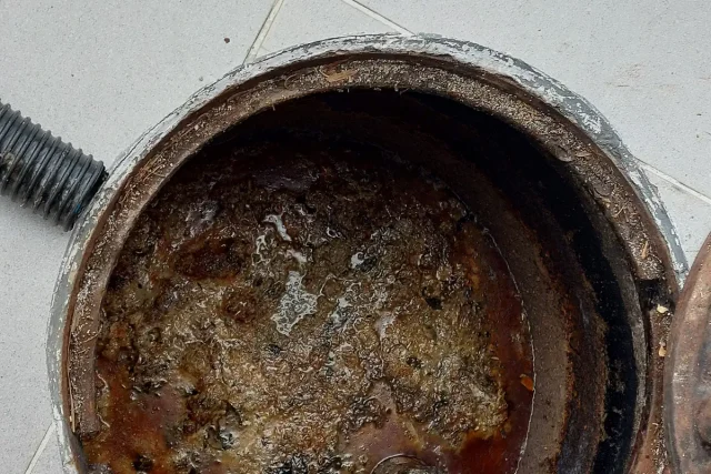 Caixa de gordura redonda vista de cima, com a borda enferrujada, mangueira preta ao lado e gordura espumosa acumulada no fundo