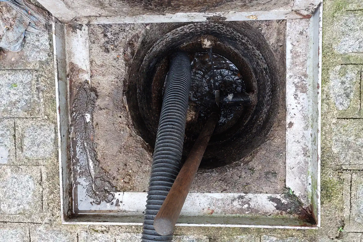 Caixa de inspeção de esgoto aberta no piso de pedras da calçada, vista de cima, com mangueira preta e haste de desentupimento dentro
