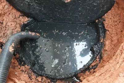 Caixa de esgoto aberta em terreno de terra vermelha, com água escura e gordurosa e uma mangueira de sucção dentro