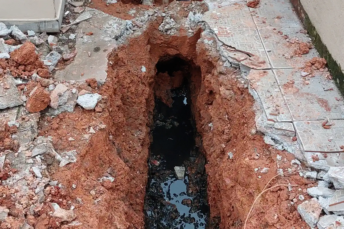Esgoto a Céu Aberto: É Crime? Onde Reclamar e Como Resolver 1 Vala aberta no quintal, com piso quebrado e terra vermelha, mostrando a tubulação de esgoto enterrada com água escura no fundo