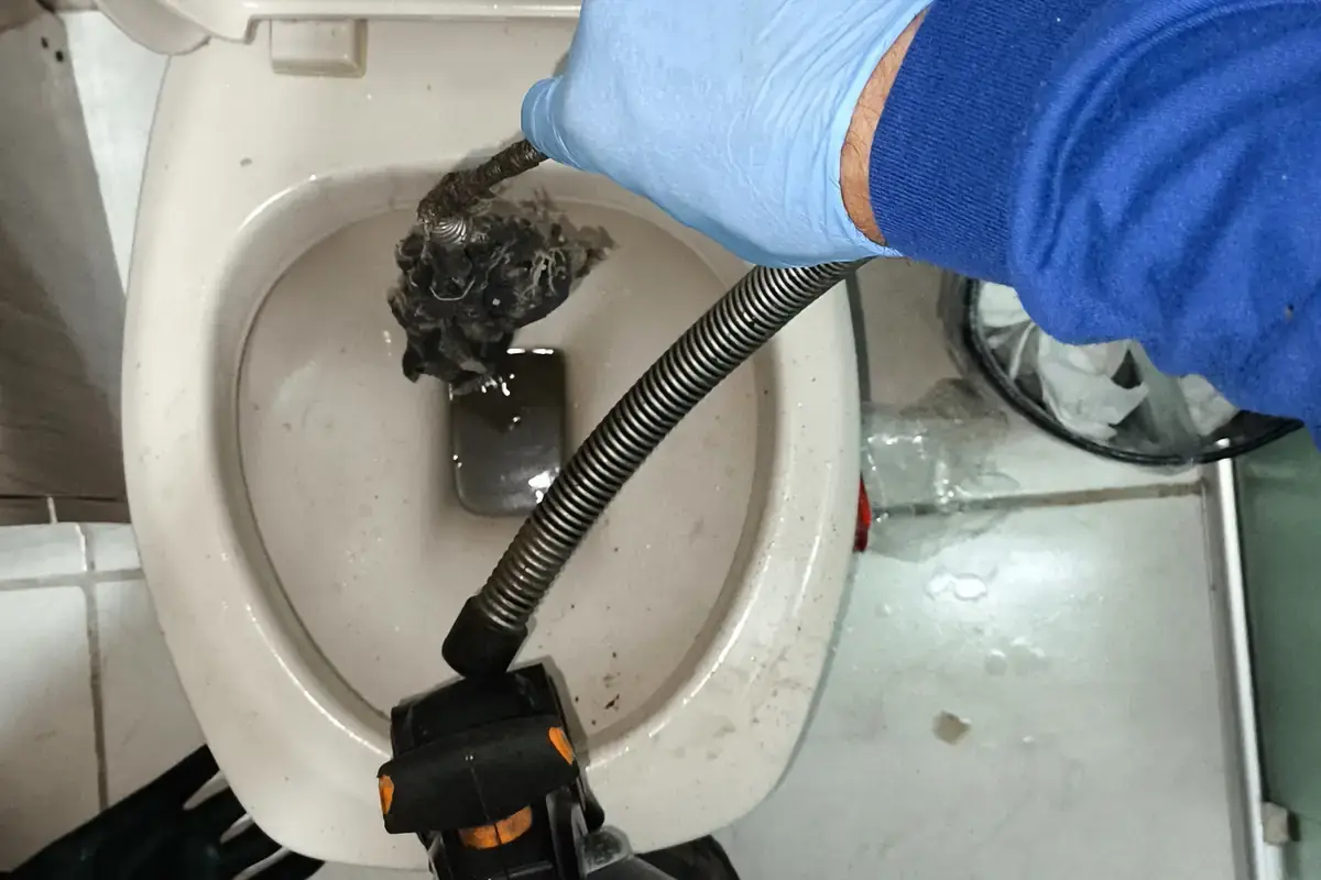 Mão com luva azul retirando um bloco de resíduos de dentro do vaso sanitário com uma mola de desentupimento