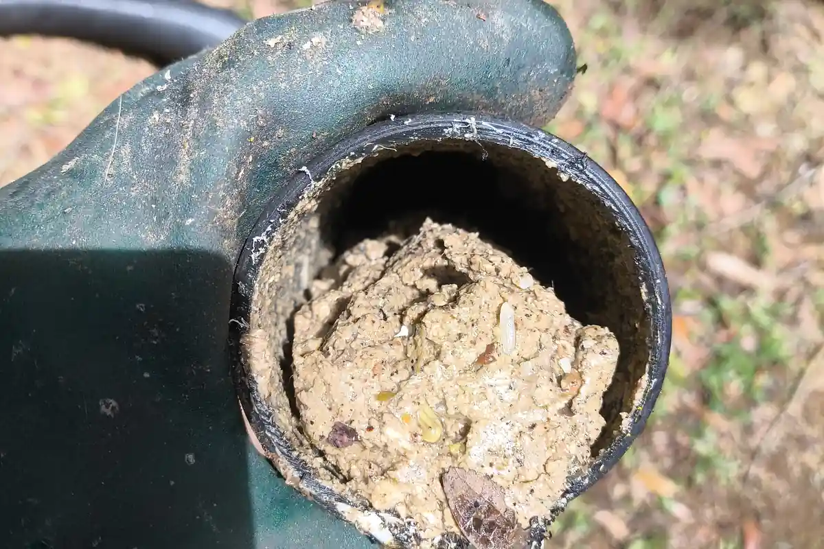 Mão com luva verde segurando uma ponta de tubo de esgoto cheia por dentro de massa de terra e resíduos endurecidos