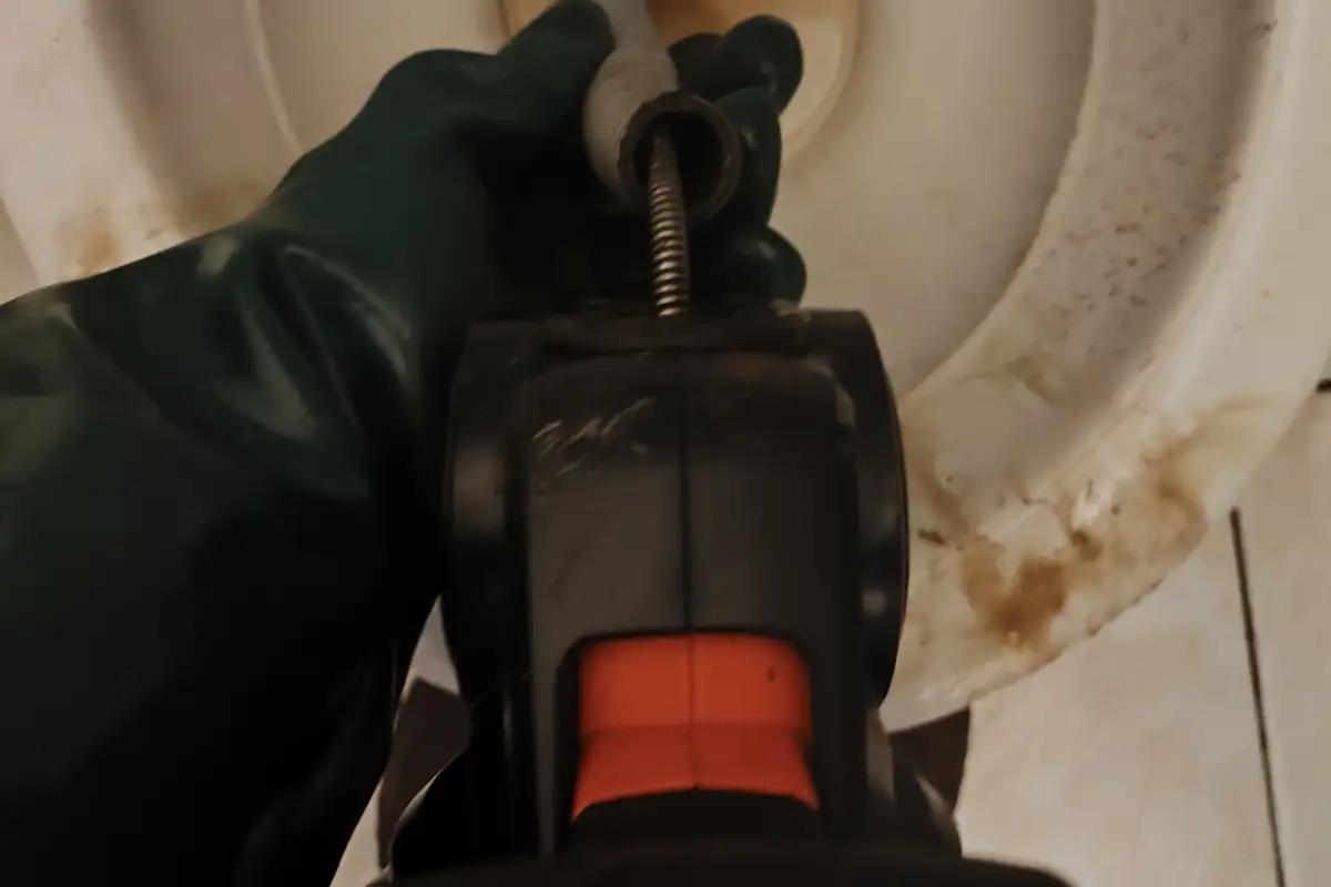 Mão com luva verde segurando a máquina de desentupimento manual com a mola dentro da bacia do vaso sanitário
