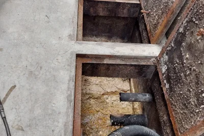 Duas câmaras de uma caixa de gordura de concreto abertas no piso, com tampa enferrujada à direita e mangueira de sucção preta na parte de baixo