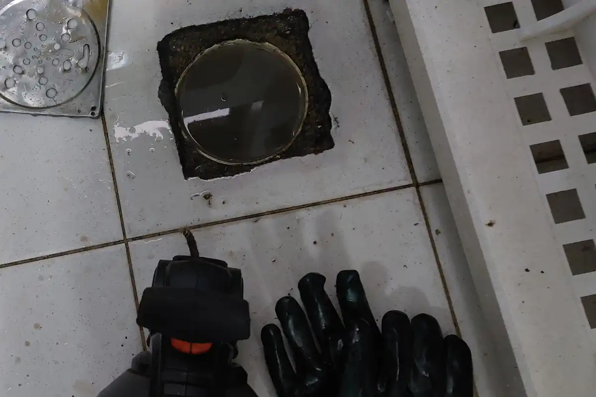 Caixa de Gordura para Restaurante: Norma, Tamanho e Limpeza 1 Ralo de piso de cozinha industrial com gordura nas bordas, máquina de desentupir preta e luvas verdes sobre o piso de azulejo branco