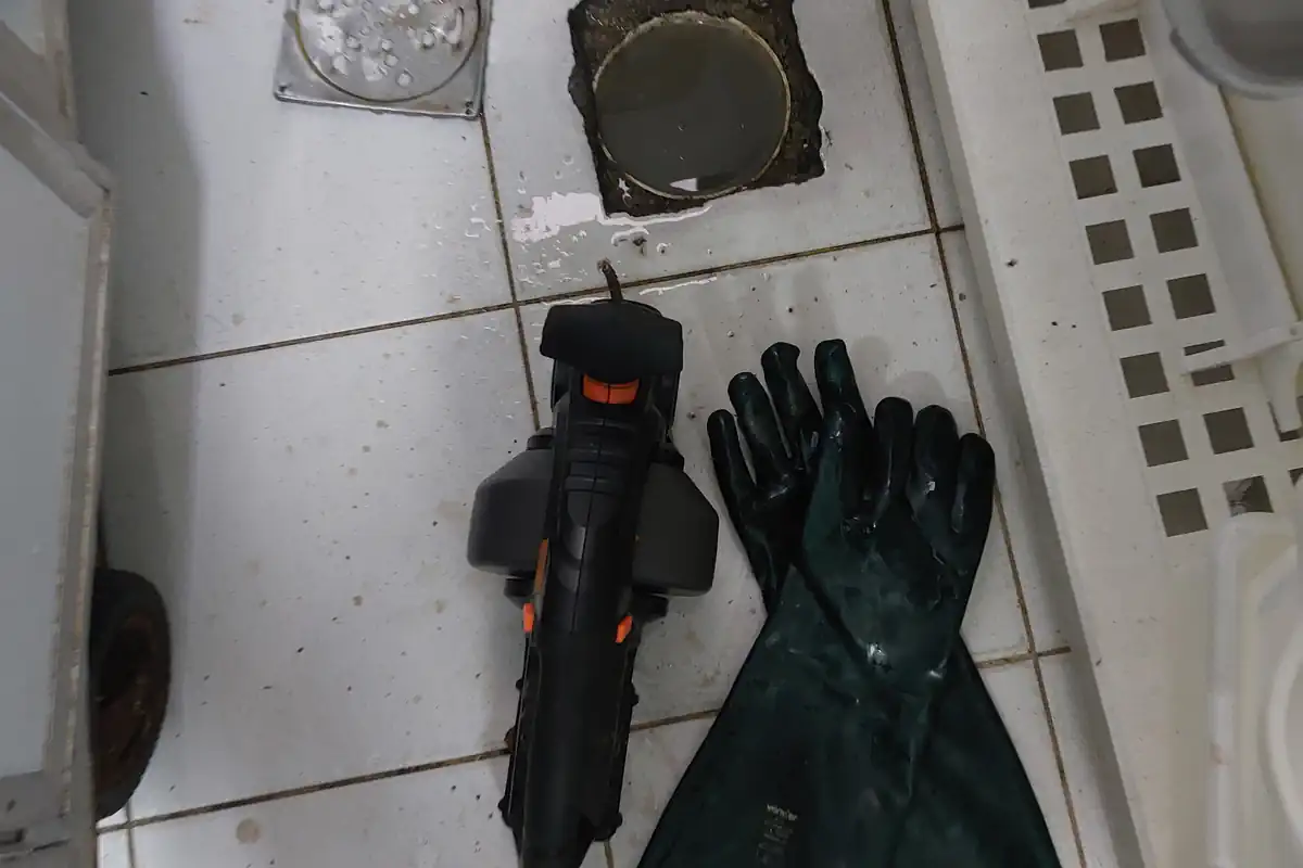 Ralo de piso de cozinha industrial com tampa metálica ao lado, máquina de desentupir preta e luvas verdes sobre o piso de azulejo branco