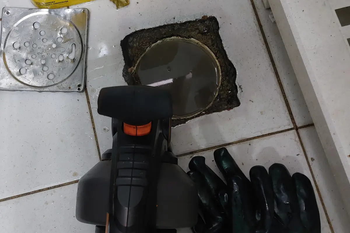 Ralo de piso de cozinha com a tampa retirada, a grelha metálica ao lado, uma máquina de desentupimento manual e um par de luvas pretas sobre o azulejo
