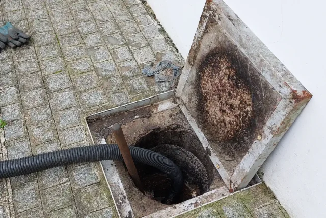 Caixa de inspeção de esgoto aberta no piso de pedras da calçada, com tampa levantada, mangueira preta, haste de desentupimento e luva ao lado